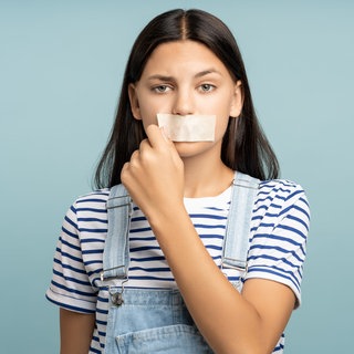 Junge Frau reißt sich Pflaster vom Mund: Tabus sind oft negativ behaftet. Veraltete Gebote, denen wir uns unterordnen. Sie ziehen aber auch Grenzen, die nicht verhandelbar sind. Der Schutz von Kindern etwa.