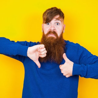 Junger Mann mit Bart zeigt "Daumen runter" und "Daumen rauf": Gutes Feedback kann Menschen sowohl im Beruf als auch im Privatleben weiterbringen. Doch wie gelingt Feedback und wie nimmt man Kritik an?