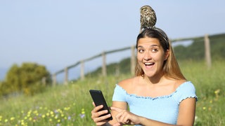 Eine erstaunte Frau, die gerade ihr Smartphone benutzt mit einem Eulenküken, das sich auf ihrem Kopf niedergelassen hat.