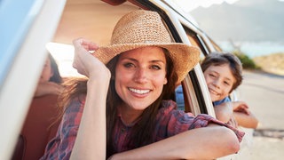 Frau mit Hut und Junge schauen aus den Fenstern eines Autos: Bei offenem Fenster steigt der Energiebedarf mit wachsender Geschwindigkeit, bei der Klimaanlage sinkt er. Wann ist also was am günstigsten?