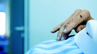 Hand eines alten Menschen auf einer Bettdecke: Selten kommt der Tod plötzlich. Er braucht Zeit. Die einen sterben schneller, die anderen langsamer. Wie unterscheiden sich klinischer Tod und Hirntod? Und wachsen Haare auch nach dem Tod noch weiter? 