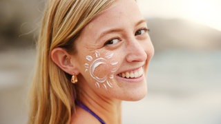 Junge Frau mit Sonnencreme in Form einer Sonne auf der Wange lacht in die Kamera: Die Haut ist unser größtes Organ. Wie bleibt meine Haut gesund und was kann ich dafür tun?