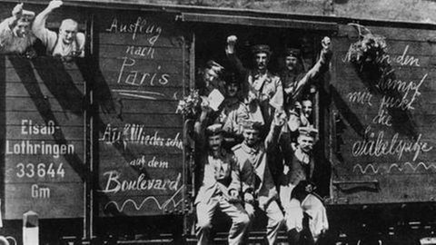 Soldaten in einem offenen Eisenbahnwaggon, winken in die Kamera, Aufschriften auf dem Waggon.