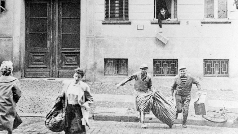 Flucht einer ganzen Familie am 20. August 1961, eine Woche nach dem Bau der Berliner Mauer. Im Hintergrund lässt ein Mann ein Paket aus einem Fenster eines Hauses in der Bernauer Straße fallen, das jetzt die Grenze markiert. Der Bürgersteig ist West-Berlin. Die Wohnungen in den Häusern wurden am 13. August evakuiert. Aber in der Anfangszeit der Mauer war die Bernauer Straße der einfachste Fluchtweg, bis die DDR alle Türen und Fenster mit Ziegeln versiegelte.