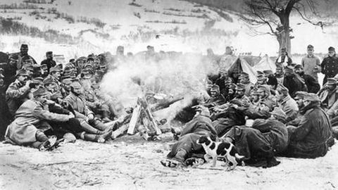 Soldaten sitzen um ein Lagerfeuer herum und wärmen sich auf, Schneelandschaft im Hintergrund.