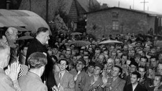 Kriegsheimkehrer hören der Begrüßungsrede von dem Minister für das Flüchtlingswesen Hans Lukaschek (2.v.l. im Profil)zu.