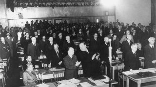 Gründung der Bundesrepublik Deutschland: Das Plenum des Parlamentarischen Rates nimmt in 3. Lesung am 8. Mai 1949 den revidierten Grundgesetzentwurf mit 53 gegen 12 Stimmen an. Vorne links: Max Reimann (sitzend), dahinter Heinz Renner, 3. von rechts: Carlo Schmid (stehend)