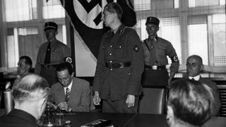 Im Haus des Beliner Rundfunks wird 1933 der neue Reichsrundfunkdirektor Eugen Hadamowsky durch Minster Joseph Goebbels eingeführt. Im Bild zu sehen ist Hadamowsky stehend bei seiner Eröffnungsrede. Links neben ihm sitzt Joseph Goebbels. Im Hintergrund zwei SA Angehörige.
