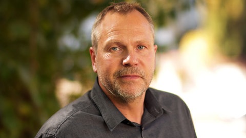 Alex Hofmann schaut in die Kamera und trägt ein graues Hemd