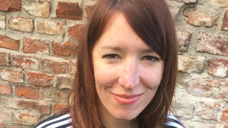 Kopf einer dunkelhaarigen Frau, sie steht vor einer Backsteinmauer