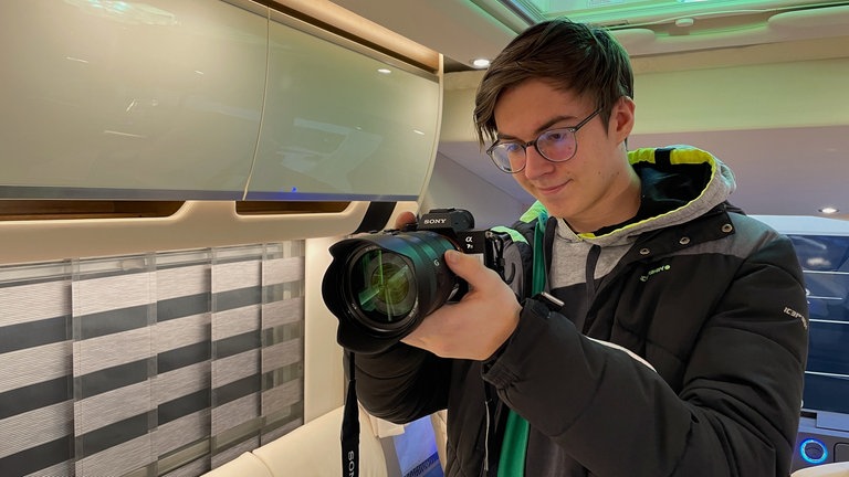 Ein junger Mann mit Brille steht in einem ausgebauten Van und hält eine Kamere in den Händen.