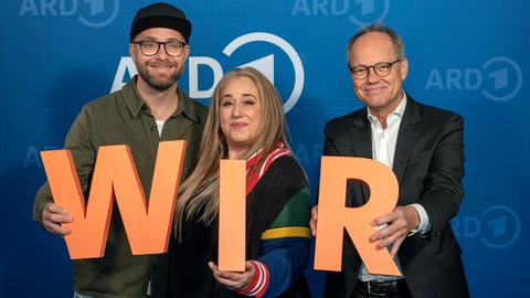 Musiker Mark Forster, Comedienne Idil Baydar und SWR Intendant Kai Gniffke (v.l.n.r.) mit orangenen Buchstaben "W I R" in den Händen, vor blauem ARD-Hintergrund