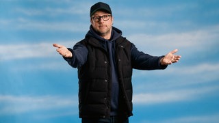 Der Schauspieler Bjarne Mädel in dunkelblauen Hoodie und Baseball Cap vor himmelblauem Hintergrund breitet die Arme einladend aus und hat einen fragenden und zugleich verschmitzten Blick