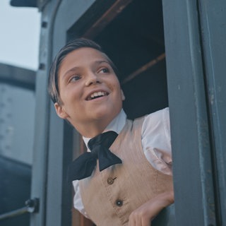 Thomas Alva Edison (Emile Cherif) beugt sich aus dem Fenster des fahrenden Zugs und lächelt.