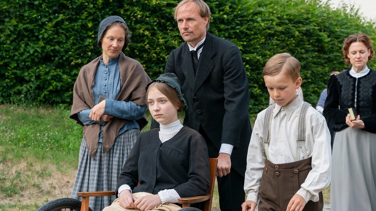 Margarete (Martha Haberland, mi.) und ihre Familie (Tessa Mittelstaedt, li.; Wolfgang Vogler, mi.; Franz Röbig, re.) laufen nach dem Kirchbesuch auf der Straße. Alle schauen etwas bedrückt.