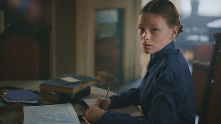 Mania (Nelly Hoffmann) sitzt am Tisch, mit einem Stift in der Hand und einem Blatt vor sich.