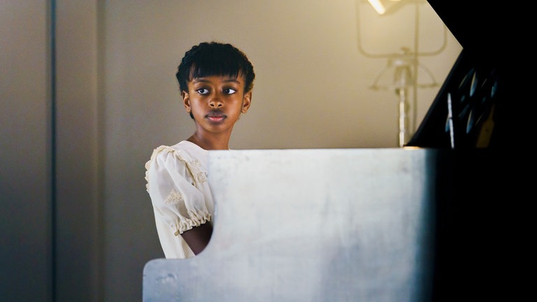 Eunice (Mahlet Paulke) sitzt am Klavier und schaut an der Kamera vorbei in Richtung Publikum.