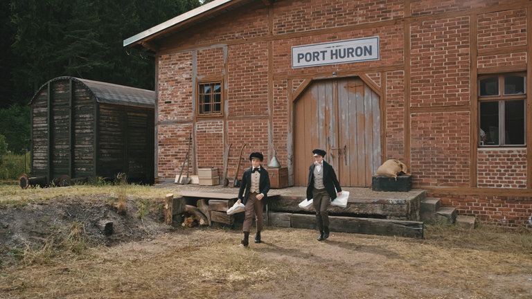 James (Ilja Bultmann, re.) und Alva (Emile Cherif, li.) laufen aus einem Gebäude mit dem Titel "Port Huron". In den Händen haben sie kleine Säckchen.