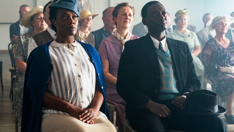 Die Eltern von Eunice Kate (Denise M'Baye, li.) und John Waymon (David Wurawa, re.) beobachten ihre Tochter bei ihrem ersten Konzert. Beide schauen angespannt an der Kamera vorbei in Richtung Bühne.