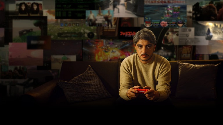 Host Khesrau Behroz sitzt mit Videospiel-Controller auf dem Sofa