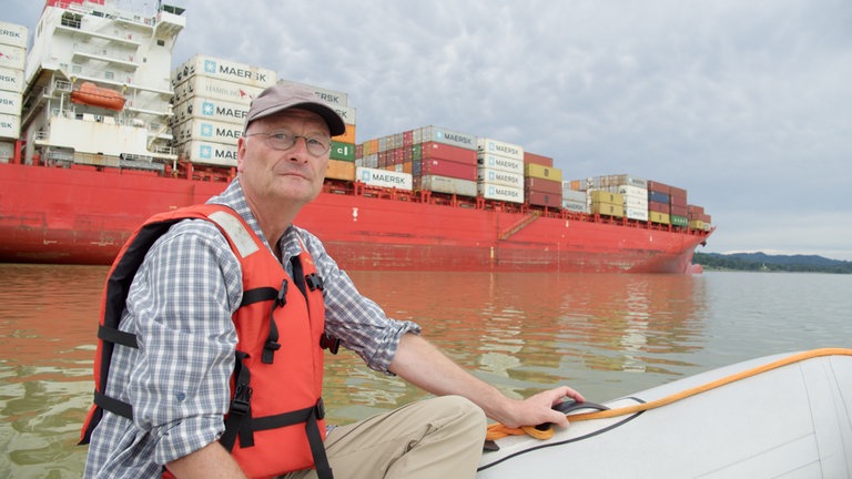 Sven Plöger auf einem kleinen Schlauchboot, dahinter ein großes Container-Schiff.