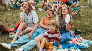 Robert Müller (Max Hubacher) hat seine Familie (Stella Goritzki, Elja Bohn, Joan Fels) zur Flugschau ausgeführt und erfüllt damit seinem Sohn einen Geburtstagswunsch. Die Familie sitzt auf einer Wiese und schaut in den Himmel. 