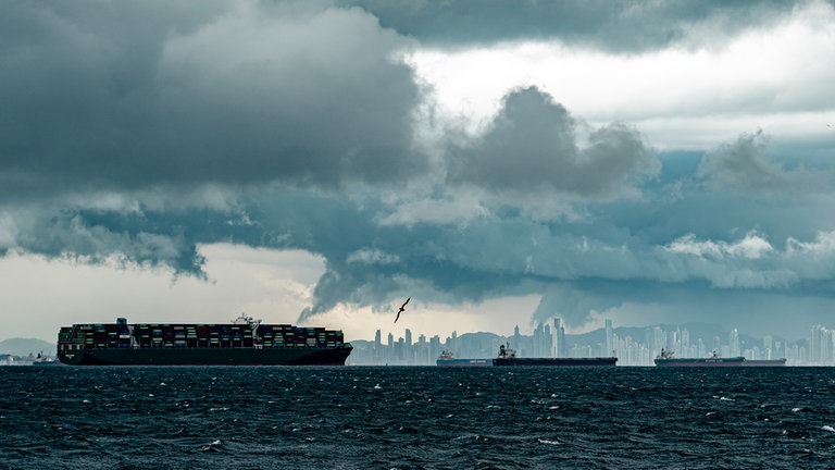Schiffe im Wasser, dahinter eine Skyline. Am Himmel dunkle Wolken.