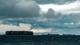 Schiffe im Wasser, dahinter eine Skyline. Am Himmel dunkle Wolken.
