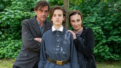 Im Foto (v.l.n.r.): Rudolf Nurejew (Mitte, Cooper Dillon), mit Vater Hamet (li., Martin Feifel) und Mutter Farida (Katerina Medvedeva) stehen vor einem Baum, im Hintergrund sind grüne Hecken zu sehen