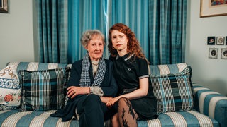 Luise (re.) und ihre Oma Guny sitzen nebeneinander auf der Couch und blicken ernst in die Kamera.