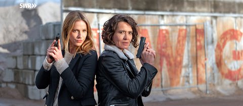 Hauptkommissarin Lena Odenthal und Hauptkommissarin Johanna Stern bilden das Tatort-Team des SWR in Ludwigshafen. Sie stehen mit gezogenen Pistolen in einem Industriegebiet.