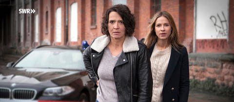 Hauptkommissarin Lena Odenthal und Hauptkommissarin Johanna Stern am Tatort. Im Vordergrund ist ein Absperrband sichtbar, während im Hintergrund der zwei Ermittlerinnen ihr Einsatzfahrzeug steht.