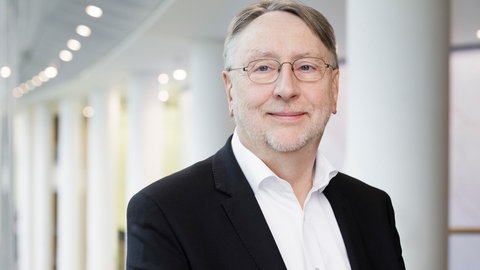 Bernd Lange, ein Mann mittleren Alters in weißem Hemd und schwarzem Sakko und Brille, lächelt dezent in die Kamera, im Hintergrund sind schlichte Betonsäulen zu erkennen