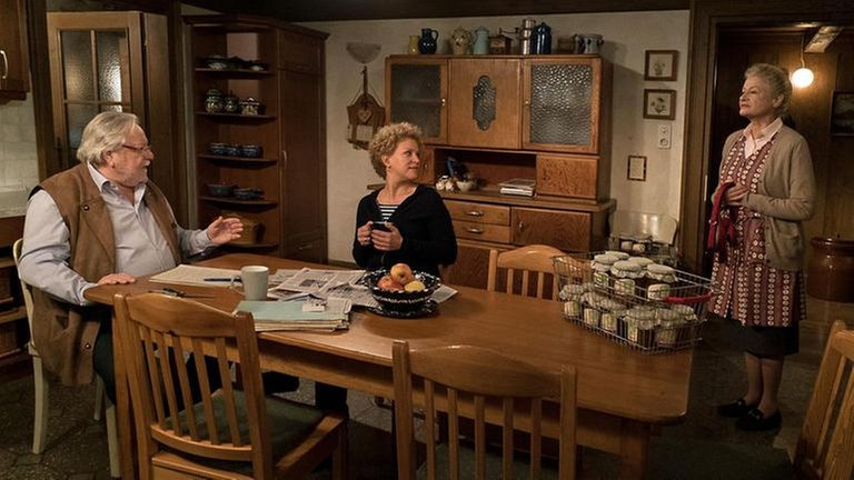 Hermann und Bea sitzen am Küchentisch, Johanna steht in der Tür und redet