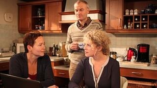 Rosi und Bea sitzen am Küchentisch, Karl steht an den Herd gelehnt neben den beiden