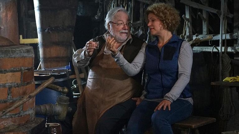 Hermann und Bea sitzen am Brennkessel und prosten sich mit Schnaps zu