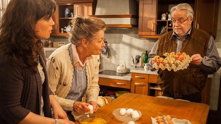 Johanna und Eva stehen am Tisch, vor sich ausgeblasene Eier, Hermann hält ein Tablett mit bemalten Ostereiern