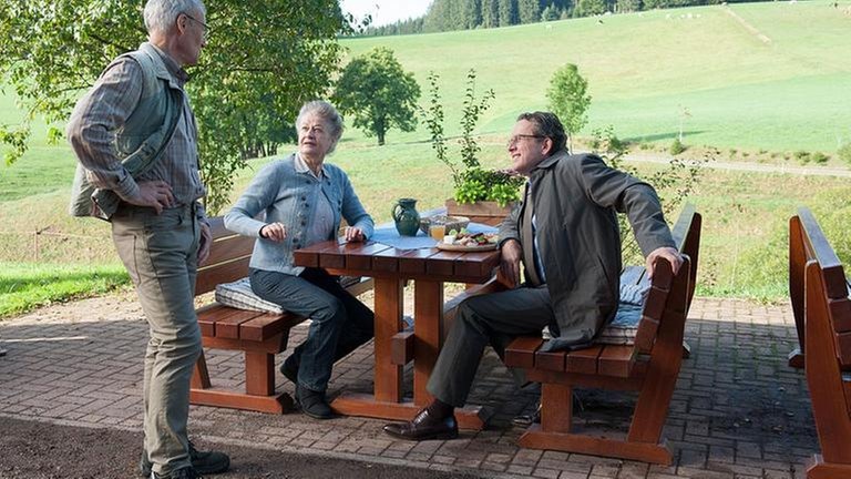 Johanna und Bernhard sitzen in der Außenwirtschaft des Hofladens, Karl steht bei den beiden