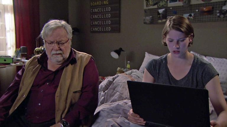 Hermann sitzt neben Celine auf dem Bett, Celine hat einen Laptop auf dem Schoß und recherchiert für Hermann