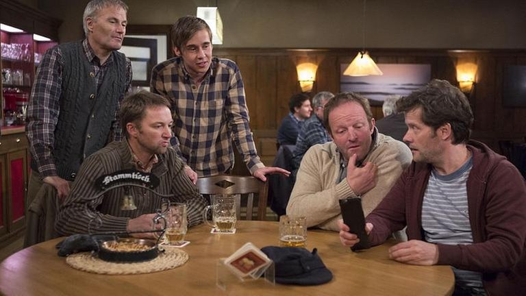 Sebastian und Karl stehen am Stammtisch, an dem Niki, Bernd und Riedle sitzen
