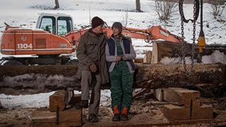Karl und Sophie sitzen im Sägewerk und unterhalten sich, im Hintergrund ein orangefarbener Bagger