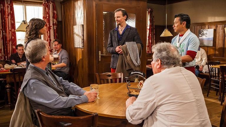 Schorsch und Leo sitzen am Stammtisch, an der Tür stehen Tu und Eva und schauen auf einen neuen Gast