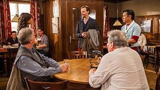 Schorsch und Leo sitzen am Stammtisch, an der Tür stehen Tu und Eva und schauen auf einen neuen Gast