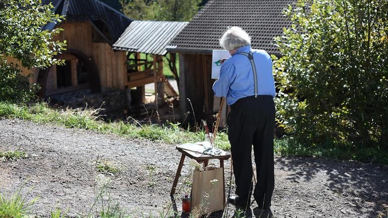 Hermann steht mit einer Staffelei vor der alten Mühle und malt