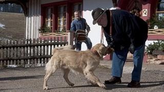 Hermann spielt vor dem Fallerhof mit Hofhund Willy, Karl kommt dazu, er trägt Stühle