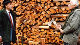 Franz und Wilhelm stehen vor einer Wand mit aufgeschichtetem Brennholz