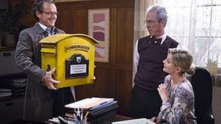 Bernhard bringt einen großen gelben Briefkasten ins Bürgermeisteramt, Frau Heilert und Herr Weiss staunen