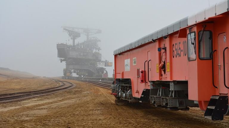 Die orange Gleisrückmaschine bereitet die Schienenanlage für den Abraumbagger im Hintergrund vor. Auch er bewegt sich auf Gleisen.