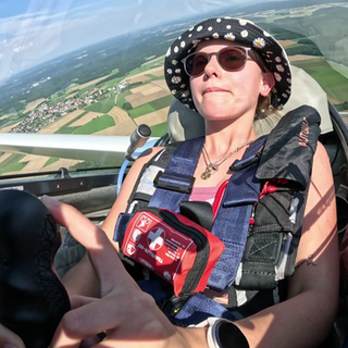 Eine junge Frau steuert ein Segelflugzeug, während im Hintergrund die Landschaft unter ihr zu sehen ist.