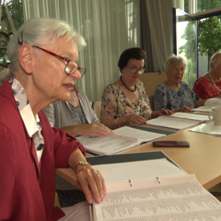 Sechs ältere Damen sitzen beisammen, halten ihre Liederbücher in den Händen und singen gemeinsam Volkslieder mit sichtbarer Freude und Konzentration.